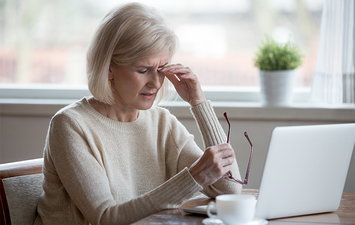 Older woman is sitting in her home, in front of a laptop. She is taking off her glasses and rubbing her left eye.