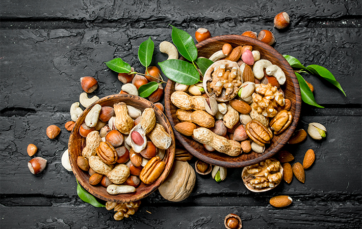 Close of a dark charcoal black wooden table with two baskets of mixed nuts on top of it. The baskets are overflowing onto the table. There is also a few green leaves scattered throughout the composition.