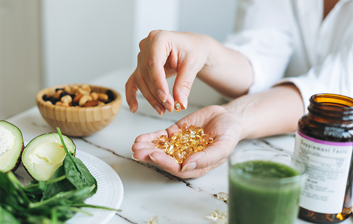 Close up of someone's hand, holding a handful of omega 3 tablets. Using the other hand to pick 1 tablet up. There are green veggies, mixed nuts, and a green smoothie surrounding the hand on a kitchen table. 