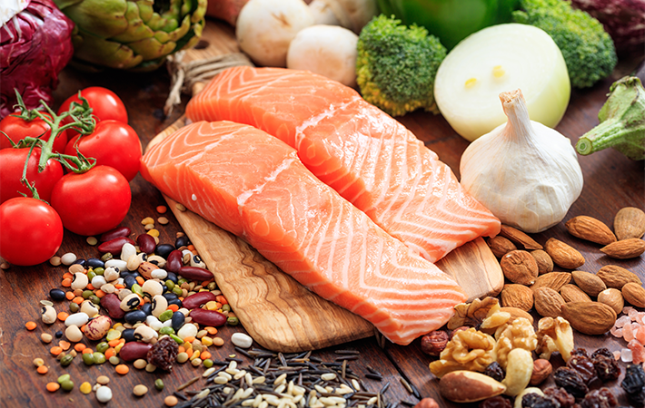 Close of a dark wooden table with food bundled on it. The main food item is 2 long cuts of pink, red salmon. There are beans, rice, nuts, tomatoes, garlic, and green vegetables surrounding the salmon.